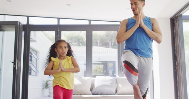 Father and young daughter practicing tree pose in a sunlit living room. Ideal for promoting family bonding, kids' exercise, healthy lifestyles, and father-daughter relationships. Suitable for wellness blogs, family-focused content, yoga class promotions, and health and fitness articles.