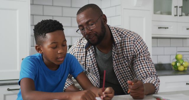 A father is assisting his son with homework in a modern, well-lit kitchen. This image can be used for educational content, family blogs, parenting articles, and advertisements promoting family-friendly products or educational tools.