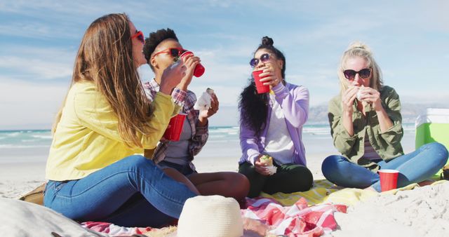 Four young women are sitting on a blanket, enjoying a beach picnic on a sunny day. They are holding drinks and eating, having a great time together. They can be wearing casual summer outfits and sunglasses, creating a vibrant and joyful atmosphere. Use this for themes such as friendship, summer activities, relaxation, or vacation promotions.
