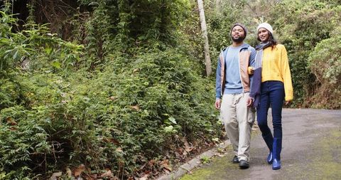 Couple linking arms walking on mossy forest path wearing knit beanies and rain boots