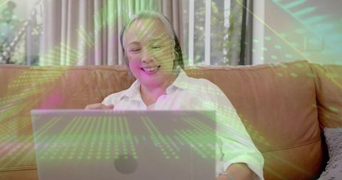 Senior woman smiling while using laptop on cozy sofa with green digital data overlay