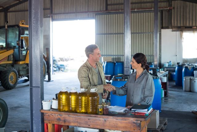 Two workers are shaking hands and smiling in an olive factory, indicating successful teamwork or a business agreement. The setting includes machinery, containers, and bottles of olive oil, suggesting an industrial and productive environment. This image can be used to represent concepts of collaboration, partnership, business agreements, and industrial production in marketing materials, business presentations, or articles about the olive oil industry.