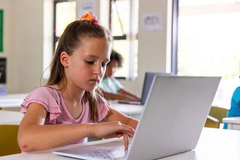 Girl typing on laptop while learning in bright classroom, focused elementary student