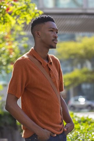 Contemplative young man standing urban green space wearing rust polo and crossbody bag