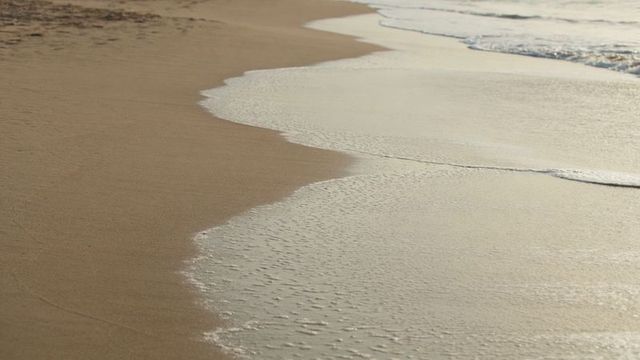 Gentle waves washing smooth sand shoreline at low tide creating scalloped foam patterns