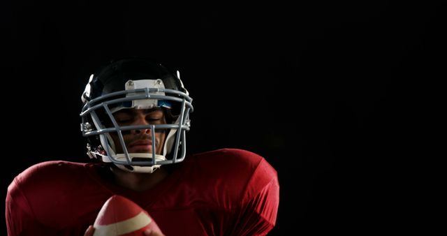 Subject is an American football player wearing helmet and padded gear, holding ball with serious expression. Dark background enhances focus on the player, showcasing determination typically seen in sports. Ideal for articles on sportsmanship, athletic training, football promotions, motivational content, or team sports advertisements.