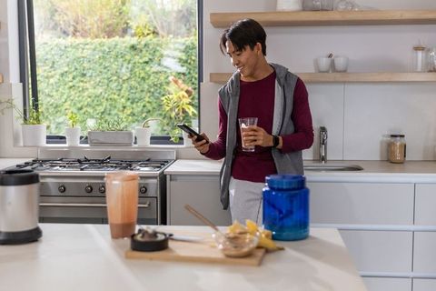 Asian man leaning in modern kitchen checking smartphone while holding morning smoothie