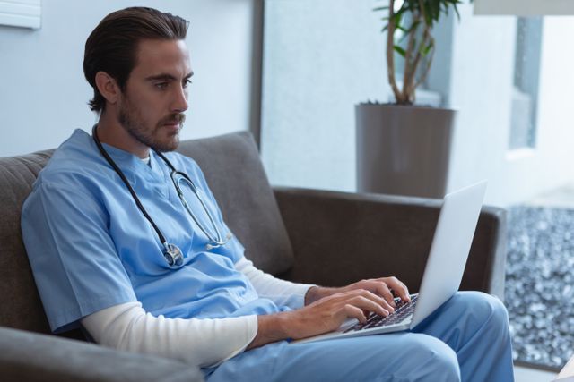 Side view of male surgeon using laptop on sofa in lobby at hospital