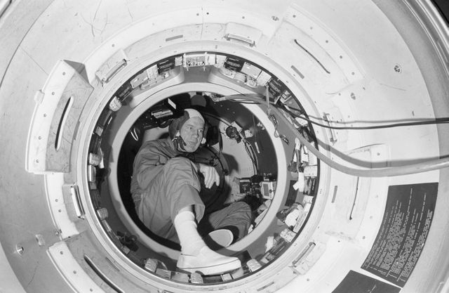 An astronaut is inside a docking module mock-up at the Johnson Space Center, training for the Apollo-Soyuz Test Project. The astronaut is sitting in a confined space surrounded by various instruments and equipment used for the mission. This scene highlights the rigorous preparation involved in space missions and the sophisticated technology used by NASA. This image can be used in articles about historic space missions, NASA training programs, and the importance of international cooperation in space exploration.