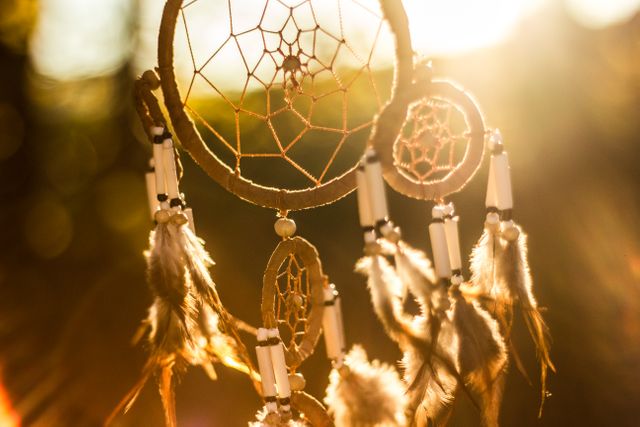 A traditional dreamcatcher adorned with feathers and beads hanging outdoors in a sunlit setting. Perfect for themes of spirituality, culture, handmade crafts, and nature. Ideal for use in articles about indigenous culture, bohemian lifestyle, spiritual practices, or as a decorative piece. The warm lighting evokes feelings of peace and serenity.