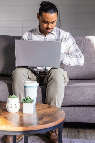 Man in thirties working on laptop on sofa in minimal home office with coffee cup