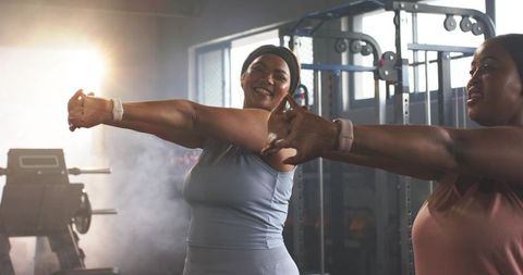 African American Women Stretching and Smiling in Gym Wearing Fitness Trackers Backlit
