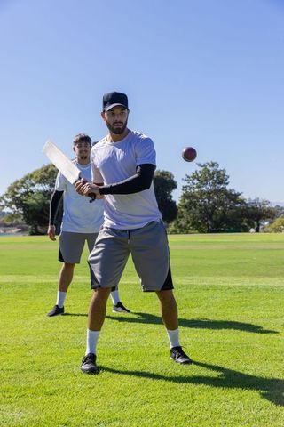 Young men practicing cricket batting on sunny park field in sports kit
