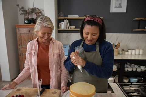 Two mature friends decorating layered sponge cake together in cozy home kitchen