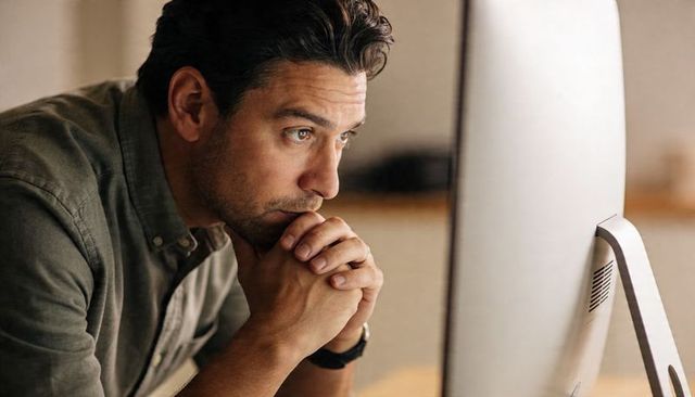 Contemplative professional leaning at desk, focusing on desktop monitor in warm light
