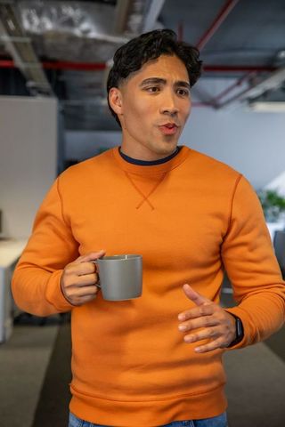 Young Asian man standing holding mug and talking in modern open-plan office, orange sweater