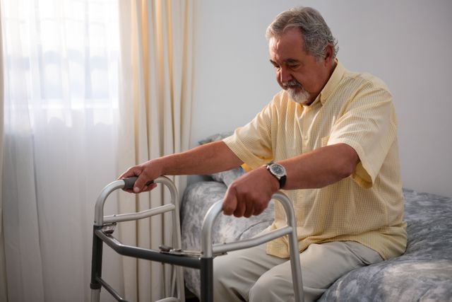 Senior man holding walker while sitting on bed in nursing home