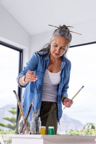 Senior woman artist selecting brushes in bright studio with jars of paint, daylight focused