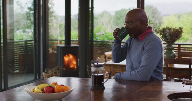 Middle-aged man sitting at wooden table drinking coffee from mug. Table features a fruit bowl and a French press coffeemaker. Large windows offer view of lush greenery while fireplace adds warmth and coziness. Perfect for illustrating rustic retreats, morning rituals, relaxation, and connection with nature.