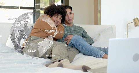 Same-sex couple cuddling on cozy bed watching laptop with augmented reality globe overlay