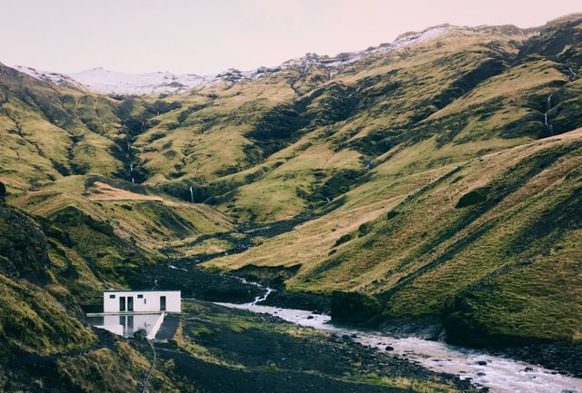 Image of a small white house situated in a remote Nordic mountain valley with green hills and a flowing river. Excellent for travel-themed projects, environmental articles, brochures on rural living or isolation, and illustrations in adventure blogs.