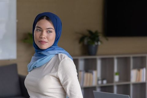 Confident woman wearing layered blue hijab standing in modern office lounge