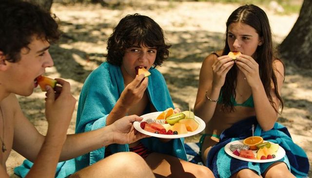 Teen trio enjoying fresh fruit picnic on sunlit sandy beach under tree shade