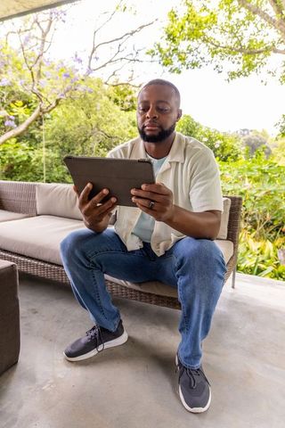 African american man reading tablet relaxing on covered patio wicker sofa in garden