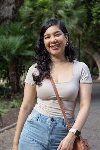 Smiling woman strolling park wearing beige top, high-waist jeans, leather crossbody bag