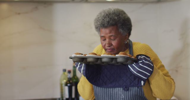 Senior woman wearing a yellow sweater and apron, holding a tray of freshly baked muffins while closing her eyes and enjoying the aroma. Oven mitts for safety, background with kitchen elements like bottles and counter. Perfect for culinary blogs, advertisements for baking products, recipes, or senior lifestyle content.
