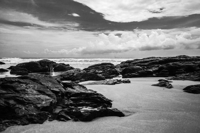 This image showcases a dramatic black and white view of a rocky beach with ocean waves crashing against the rocks and dramatic clouds covering the sky. The contrasting elements provide a timeless and calming atmosphere. This can be used for backgrounds, nature themes, or to evoke a sense of tranquility and contemplation.