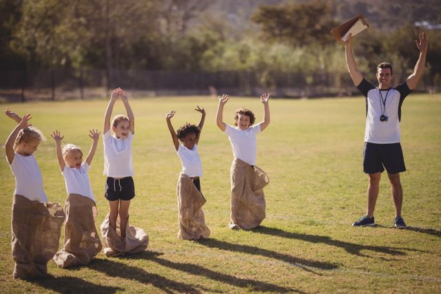 Coach and schoolgirls participating in a sack race outdoors on a sunny day. The children are raising their hands in excitement while the coach cheers them on. This image can be used for promoting outdoor activities, school events, physical education programs, and children's sports competitions.