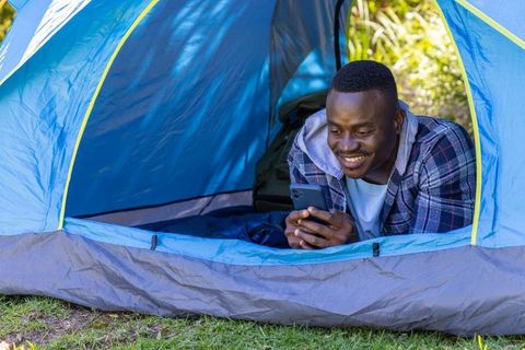 Smiling african american camper relaxing in blue tent using smartphone on green campsite
