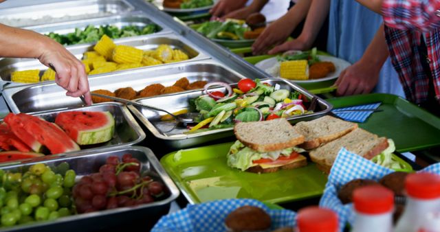 A diverse group of people is serving themselves a variety of healthy foods from a buffet line, with copy space. Fresh fruits, vegetables, and sandwiches offer a glimpse into a communal meal setting, promoting healthy eating habits.