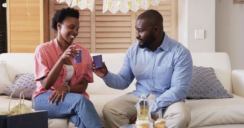 African American couple clinking cups celebrating with cupcakes on cozy living sofa