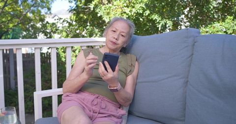 Senior asian woman relaxing on sunlit deck sofa using smartphone for staying connected