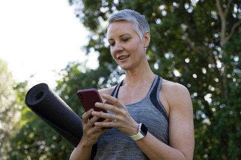 Fit middle-aged woman checking phone after outdoor yoga holding mat in park with smartwatch