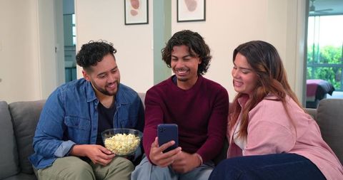 Friends watching video on smartphone while sharing popcorn on couch in relaxed living room