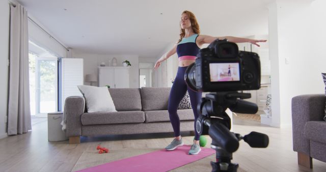 Woman practicing yoga in living room while recording video on camera. Great for content about online fitness classes, home workout routines, healthy living, and remote coaching.