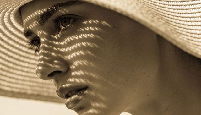 Artistic sepia close-up woman wearing wide-brim straw hat casting striped shadow across face