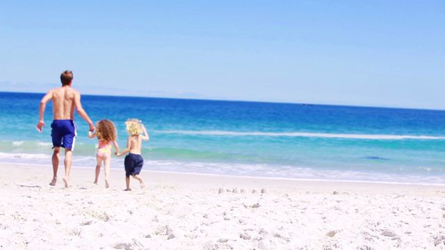 Father and his children running together on a beach. The family is enjoying a sunny day by the ocean with clear blue skies and white sand. This video is great for advertising summer vacations, family bonding activities, beach resorts, and outdoor fun.