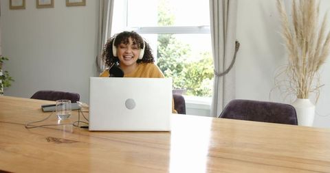 Young woman recording podcast at home dining table with laptop, microphone, headphones