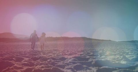 Couple walking hand in hand on rippling beach at pastel sunset with bokeh light flares