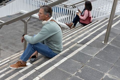 Sitting on urban stairs using smartphones, man wearing headphones and woman drinking coffee