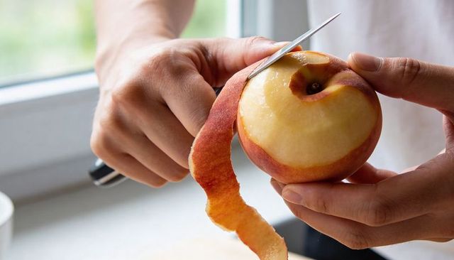 Hands peeling ripe peach with paring knife, long spiral peel falling, kitchen closeup