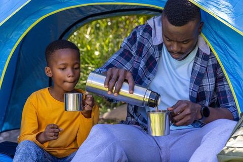 African american father and son pouring hot drink from thermos while camping in blue tent