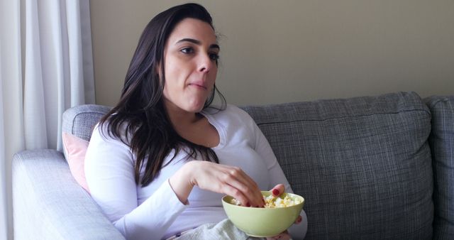 A young Caucasian woman is sitting on a couch enjoying popcorn, with copy space. Her relaxed posture and the casual setting suggest a leisurely activity, watching television or a movie.