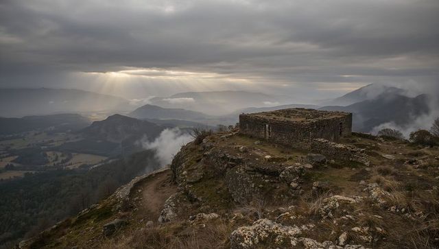 Perched stone ruin on misty mountain ridge with sunbeams piercing dramatic cloudy sky