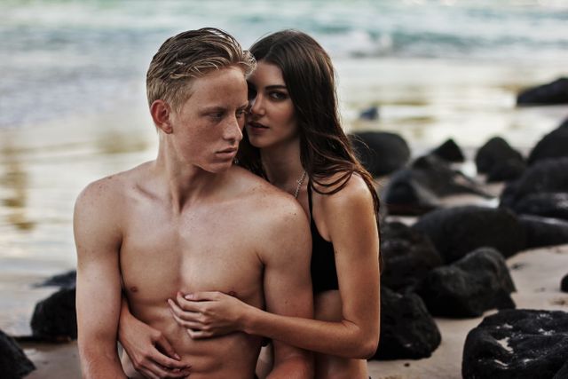 A young couple is embracing on a beach during sunset. The shirtless man and the woman are enjoying a romantic, intimate moment near the ocean with rocks in the background. Ideal for themes related to romance, love, summer vacations, beachside holidays, and carefree lifestyles.