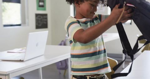 Elementary student packing backpack at classroom desk with laptop and striped shirt